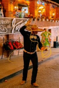 A charro spinning a lasso in the street in front of Jazmin's Restaurant, with a folkloric dancer in a green dress behind him