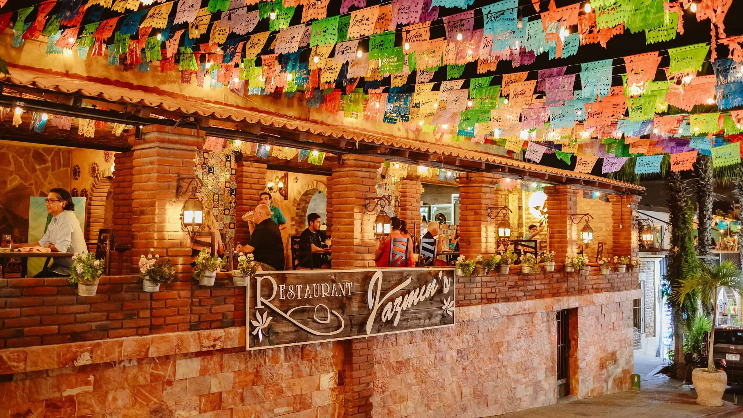 Diners on the brick terrace of Jazmin's Restaurant at night, under strings of papel picado and lanterns, above the Restaurant Jazmin's sign