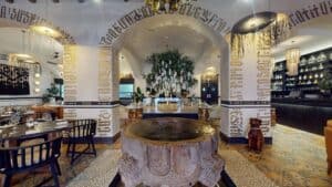 A carved stone fountain framed by arches painted with gold black-letter script inside Los Deseos, with the bar on the right