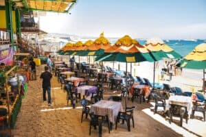 Tables with striped cloths under umbrellas on the sand at Mango Deck before the crowds arrive, with Médano Beach and boats on the bay