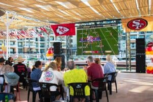 Guests at tables under the shade canopy at Mango Deck watching a football game on a giant screen