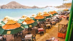 Rows of orange and green Mango Deck umbrellas over tables on the sand, facing the bay and the rocky hills of Cabo San Lucas