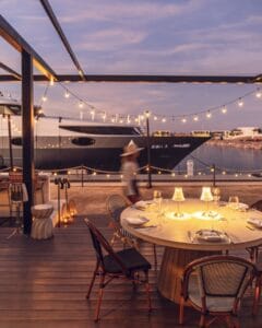 A candlelit table on the Mozza Baja deck at dusk, with string lights and yachts moored in the Costa Palmas marina