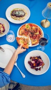 A pizza with prosciutto and chiles alongside other shared plates on a blue table at Mozza Baja