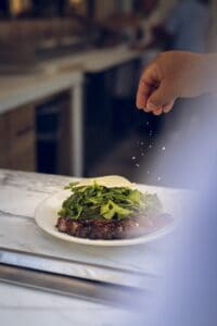 A cook seasoning a grilled steak topped with arugula and shaved Parmesan at Mozza Baja