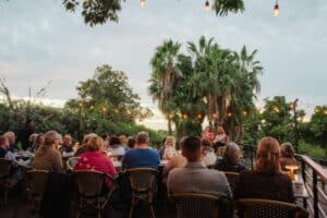 Guests at long tables on the deck terrace at Oystera at dusk, under string lights and fan palms