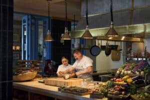 Two chefs at work in the open kitchen at Palmerio, with fresh vegetables on the counter under copper lamps