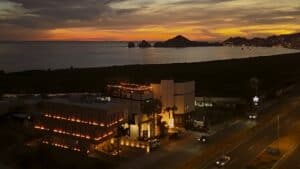 Aerial view of Puerta Vieja lit up at dusk beside the Transpeninsular Highway, with the Arch of Cabo San Lucas on the horizon
