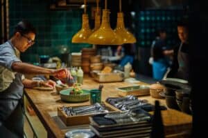 A cook plating a dish at the wooden pass of the open kitchen at Talay, under yellow pendant lamps