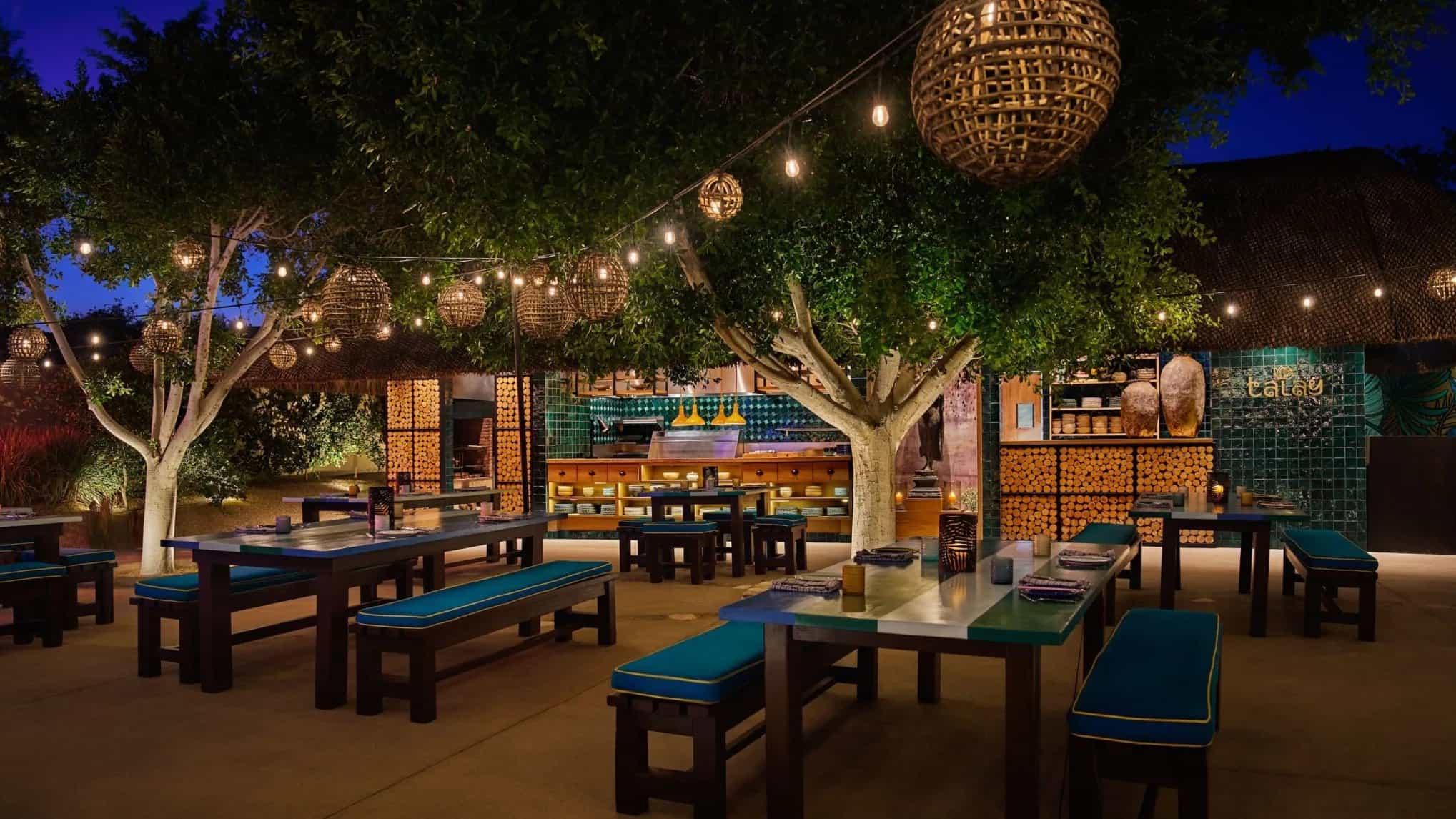 Outdoor tables and turquoise benches under trees and string lights at Talay, with the open kitchen at the back