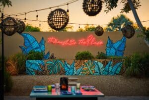 A table set in front of the tropical-leaf mural and red neon sign at Talay, under hanging woven lanterns at dusk