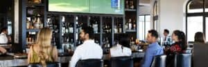 Guests on leather stools at the bar of The Woods Cabo, with golf on the big screens above the back bar
