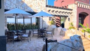 The stone-paved patio at The Woods Cabo, with iron tables under white umbrellas and the Pacific in the distance at Diamante