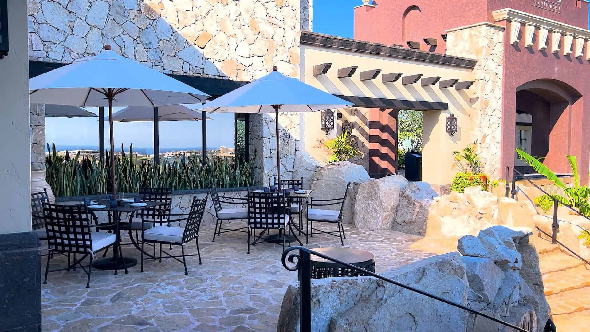 The stone-paved patio at The Woods Cabo, with iron tables under white umbrellas and the Pacific in the distance at Diamante