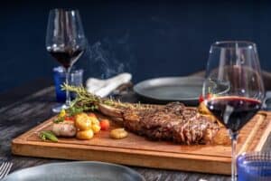 A grilled bone-in steak on a carving board with roasted potatoes, garlic and red wine at The Woods Cabo
