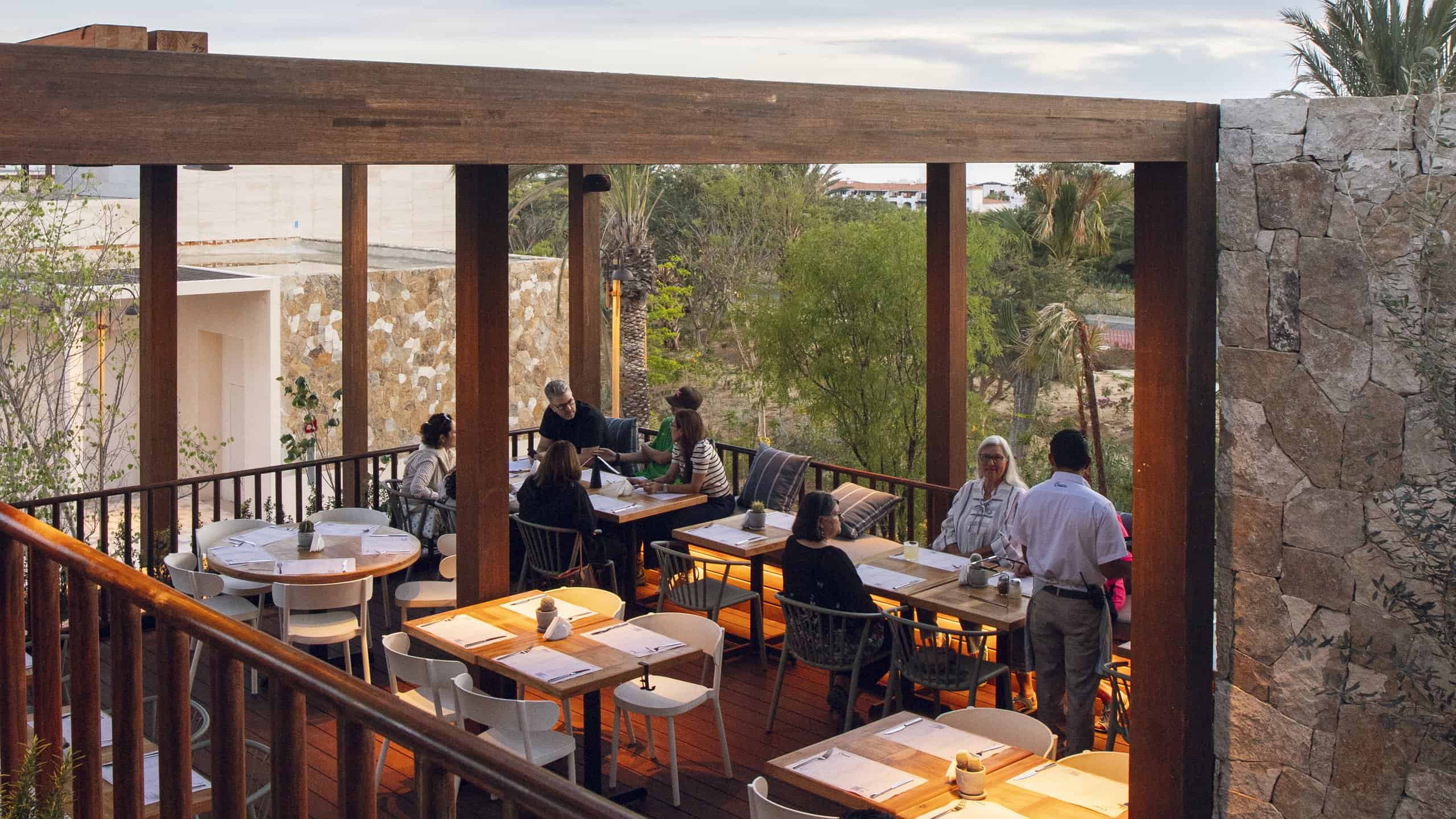 Diners at tables on the rooftop terrace of Cream Ánima Village in Cabo del Sol, under a wooden pergola in the late afternoon