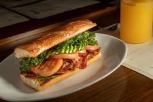 A toasted baguette sandwich with bacon, tomato, avocado and greens next to a glass of orange juice at Cream