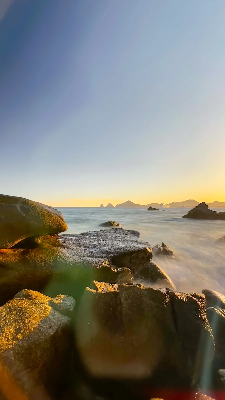 Golden evening light on granite boulders along the Cabo San Lucas shore, with the rocks of Land's End on the horizon — photo by Adrián De Antoni