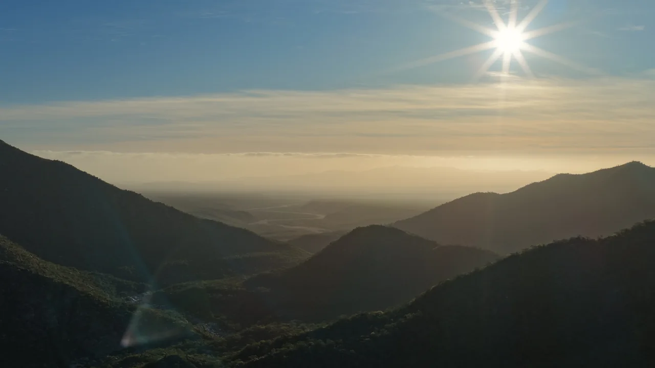 The sun bursting over the Sierra de la Laguna, with a valley opening toward the coastal plain — aerial photo by Adrián De Antoni